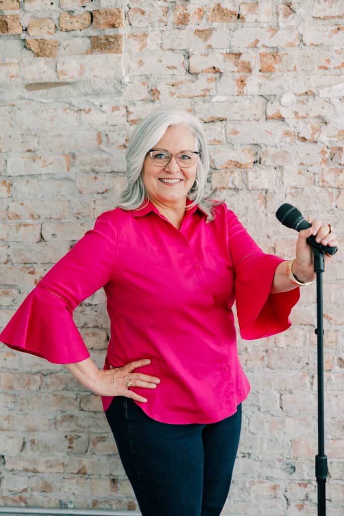 Older woman with gray hair and glasses, wearing a bright pink top and jeans, stands in front of a brick wall while holding a microphone and smiling at the camera.