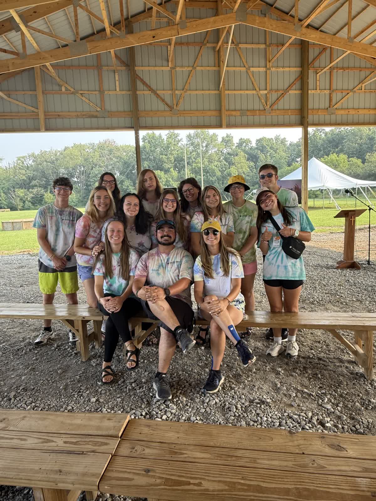 A group of thirteen people wearing tie-dye shirts poses for a photo under an open pavilion, with benches and trees visible in the background.