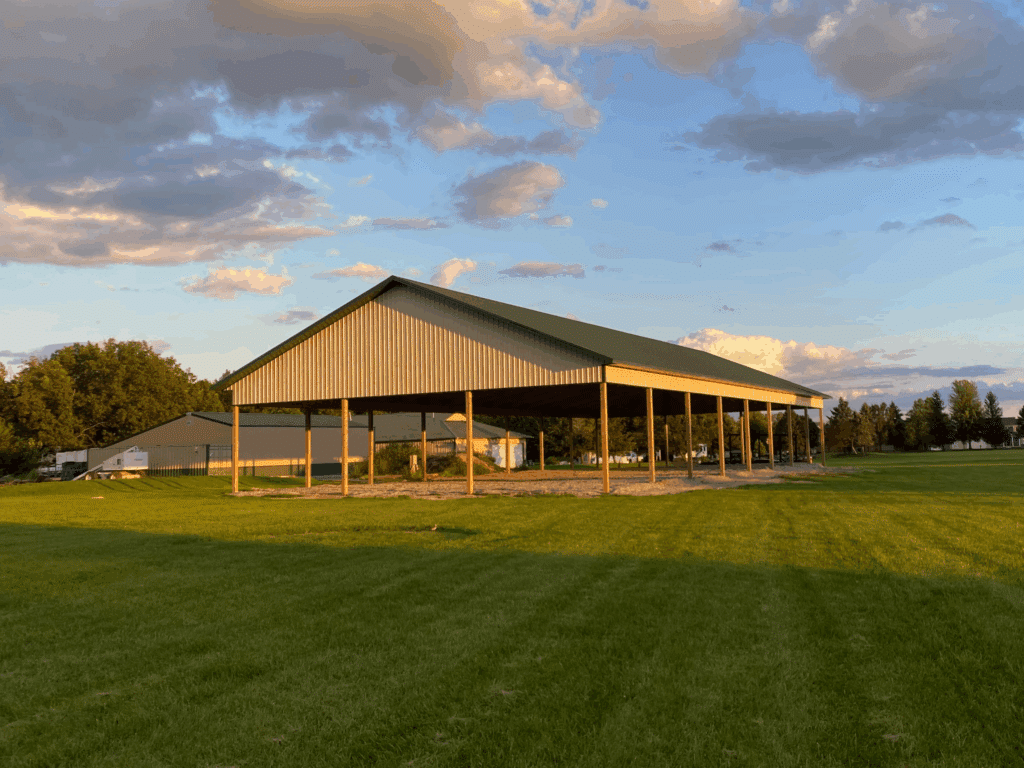 A large, open-sided metal pavilion structure stands on a grassy field under a partly cloudy sky at sunset.