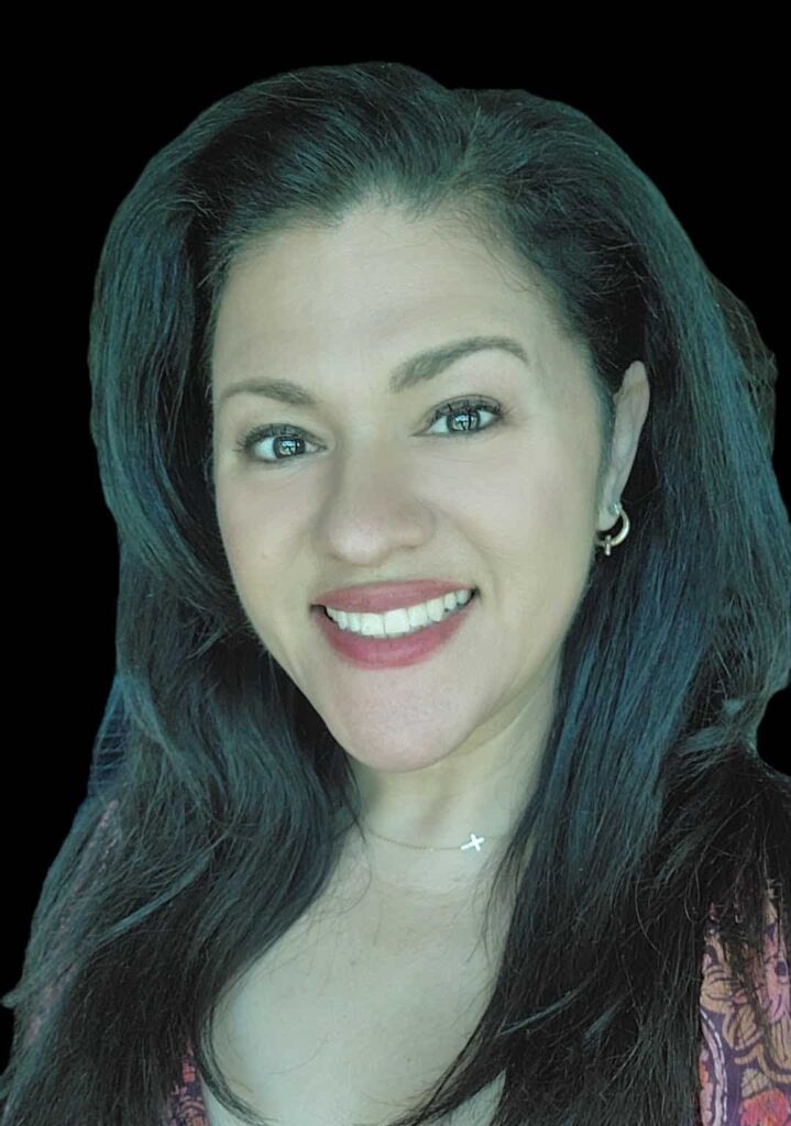 Woman with long dark hair, wearing a reddish patterned top, hoop earrings, and a star necklace, smiling at the camera against a black background.