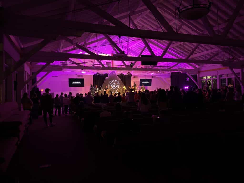 A crowd faces a stage with musicians and two screens in a dimly lit hall with purple lighting and exposed beams.