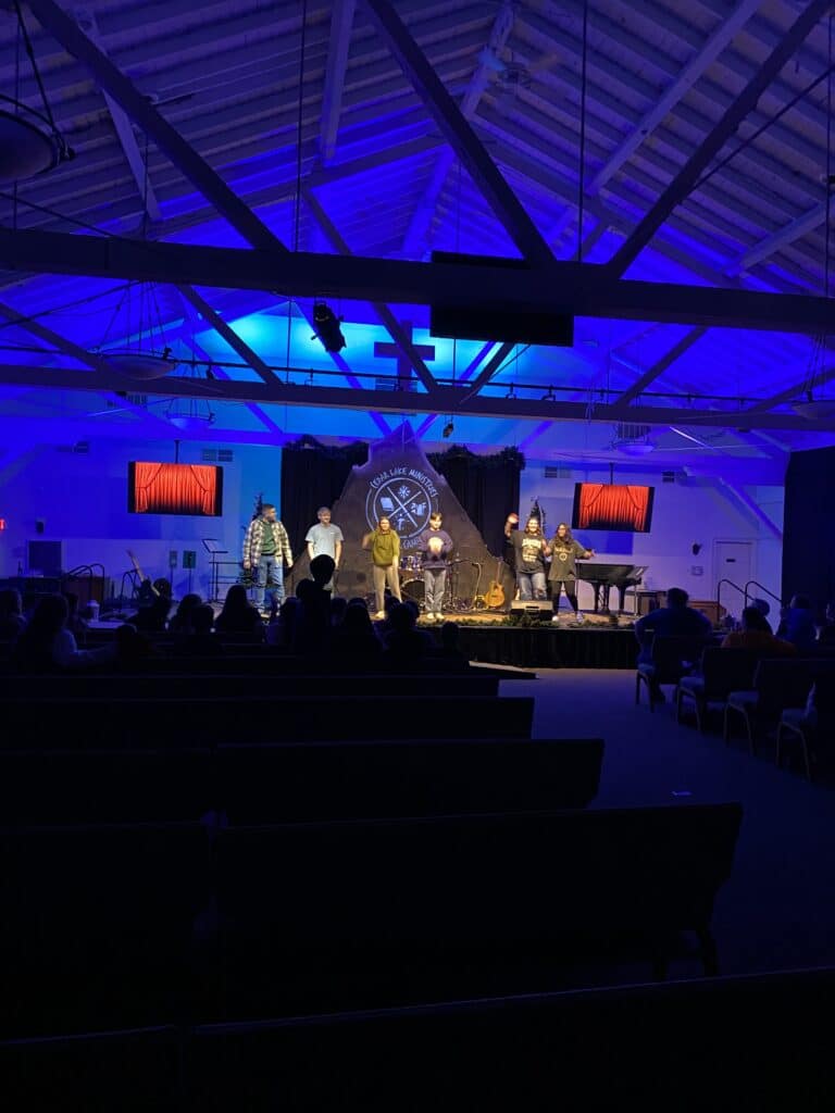 Six people stand on a stage under blue lighting, performing in front of an audience in a large room with exposed beams and a backdrop featuring a circular emblem.