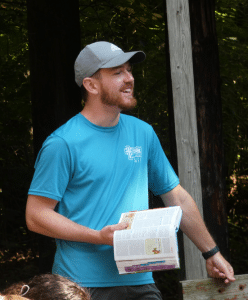 Man in a blue shirt and gray cap stands outdoors holding an open book, speaking to a group; trees and wooden beam in the background.
