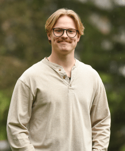 A young man with blond hair, glasses, and a mustache is standing outdoors, wearing a beige long-sleeve henley shirt. Greenery is visible in the blurred background.