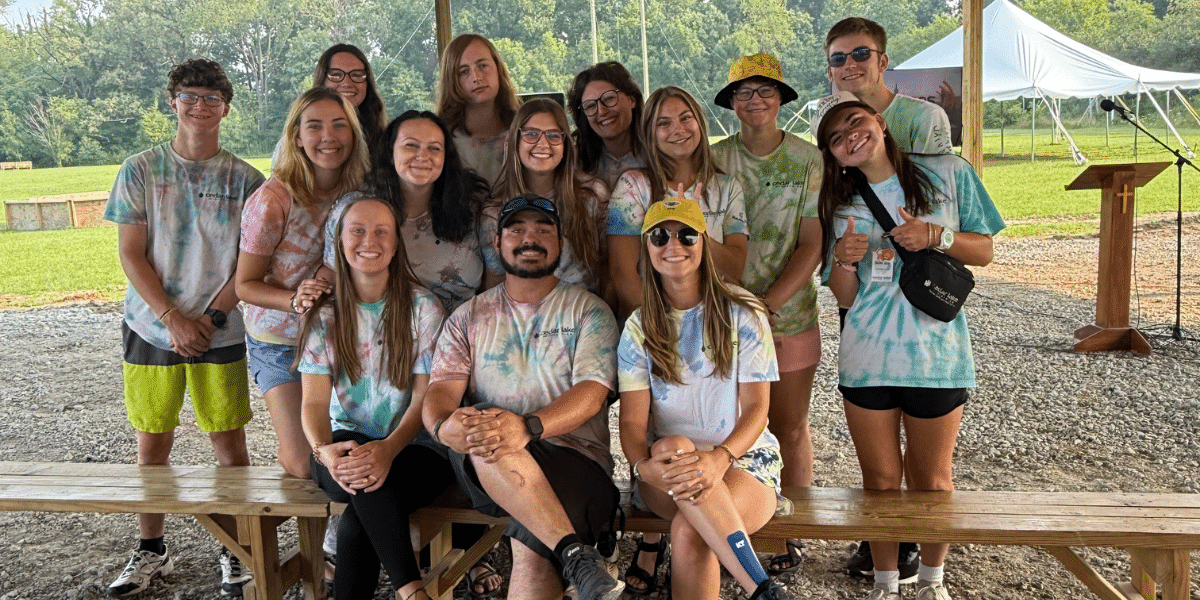 A group of people wearing tie-dye shirts pose together outdoors under a canopy, sitting and standing around wooden benches.