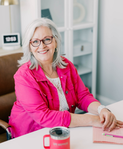 An older woman with gray hair and glasses, wearing a bright pink jacket, sits at a desk with a notebook, pen, and red mug, smiling at the camera.
