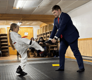 A young girl in a martial arts uniform kicks a padded target held by a man in a suit inside a room with an audience seated in the background.