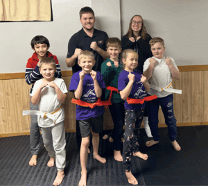 A group of children and two adults pose in a martial arts studio, some wearing colored belts and holding fighting stances, all smiling at the camera.