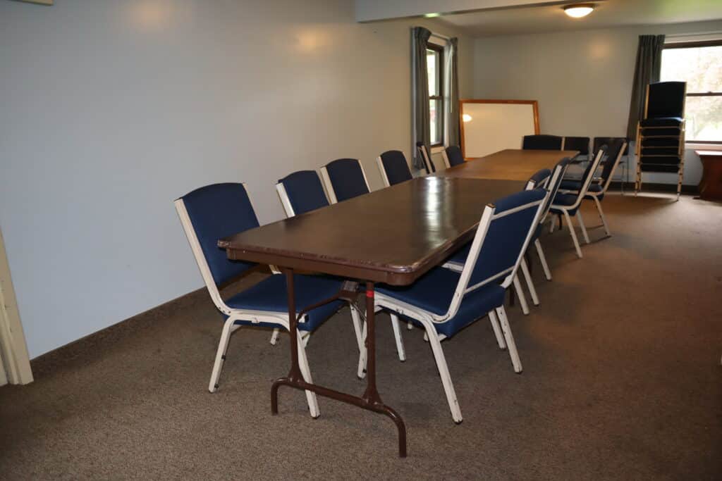 A conference room with a long brown table surrounded by blue chairs, some additional chairs stacked in the background, and windows letting in natural light.