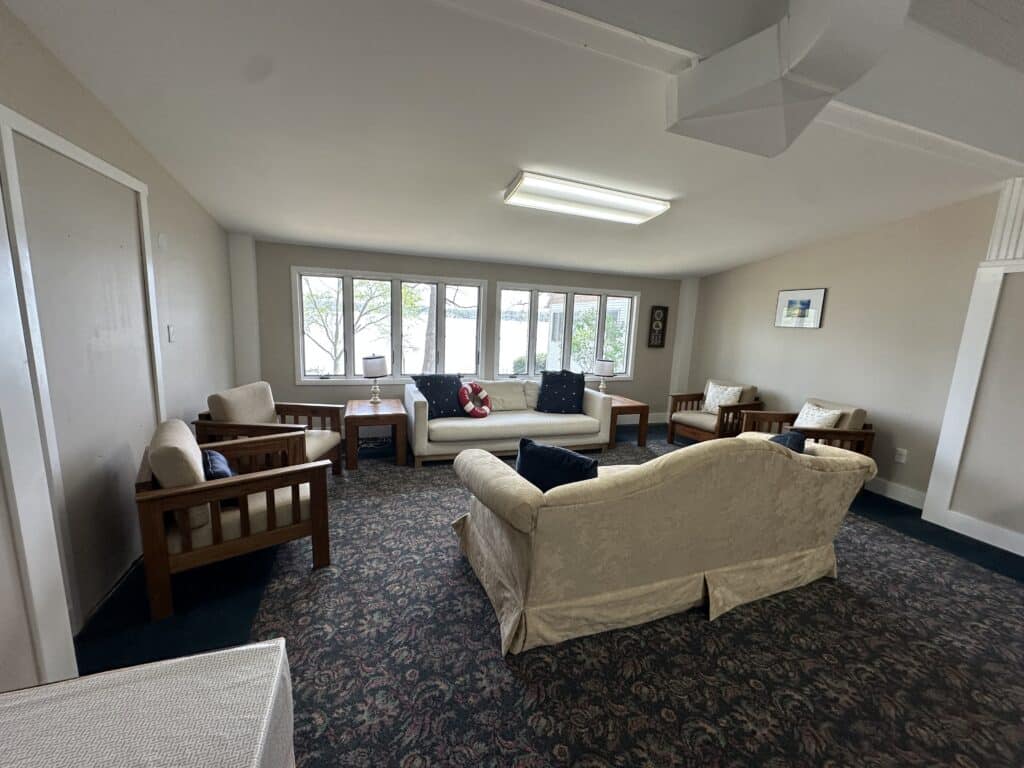 A living room with two sofas and three armchairs arranged around a small table, featuring large windows, carpeted floor, and neutral-colored walls.