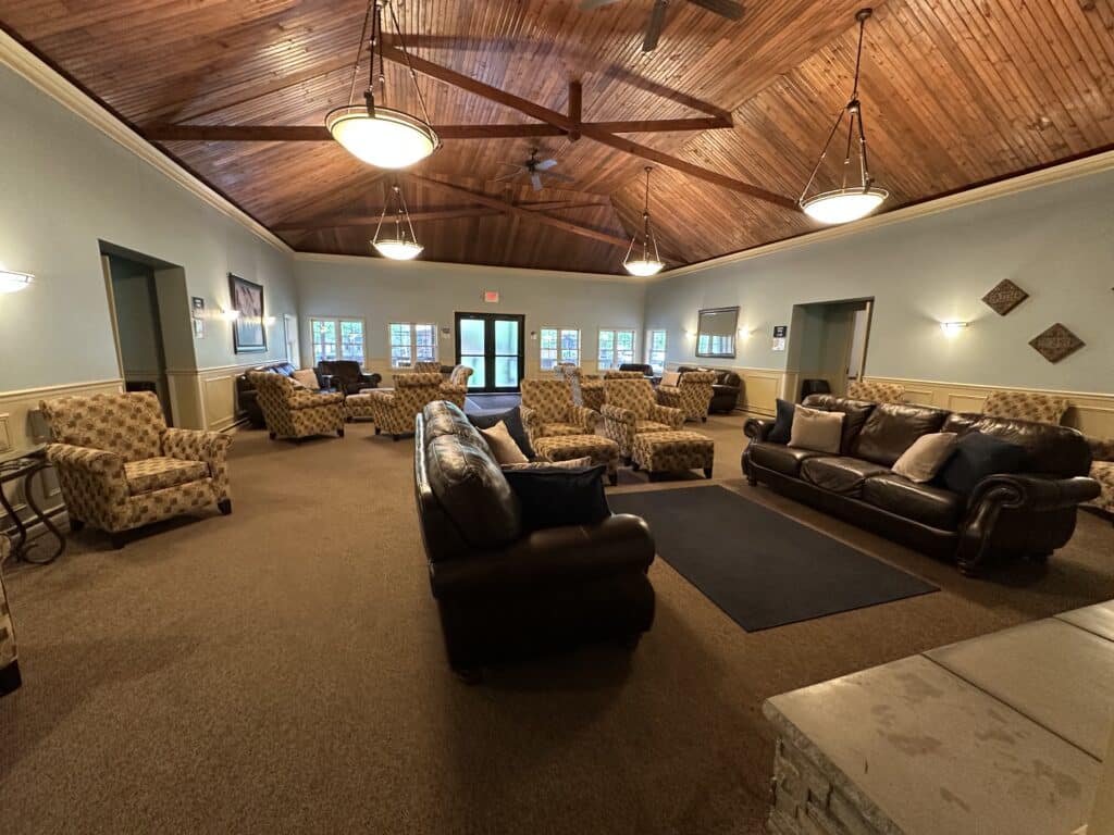 Spacious lounge with armchairs, sofas, and a vaulted wood ceiling, featuring multiple light fixtures and large windows along the far wall.