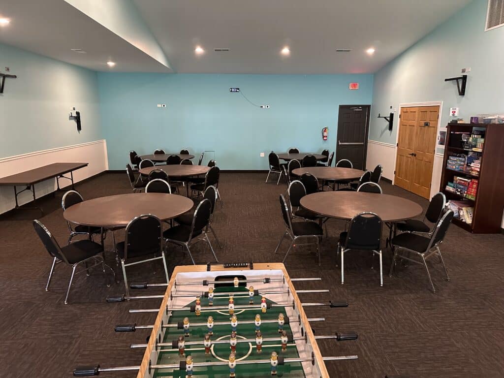A room with round tables and chairs, a foosball table in the foreground, wall-mounted TV brackets, and shelves with board games along the right wall.