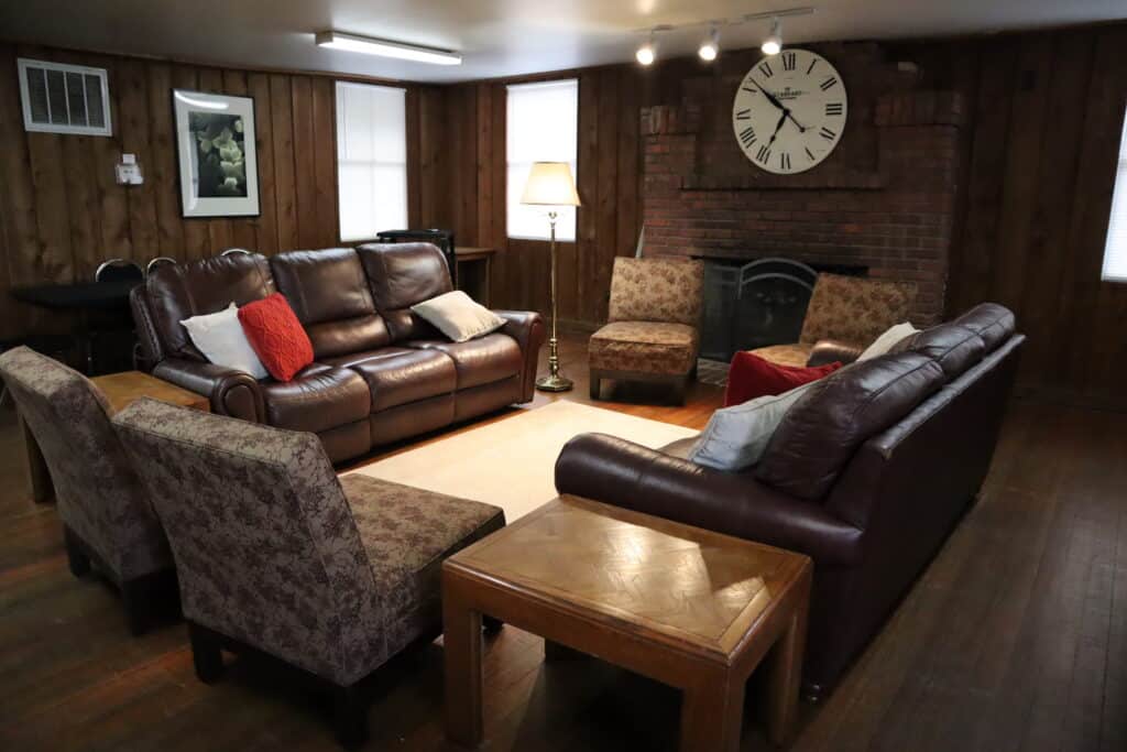 A cozy living room with two brown leather sofas, four patterned armchairs, a wooden coffee table, and a large clock above a brick fireplace.