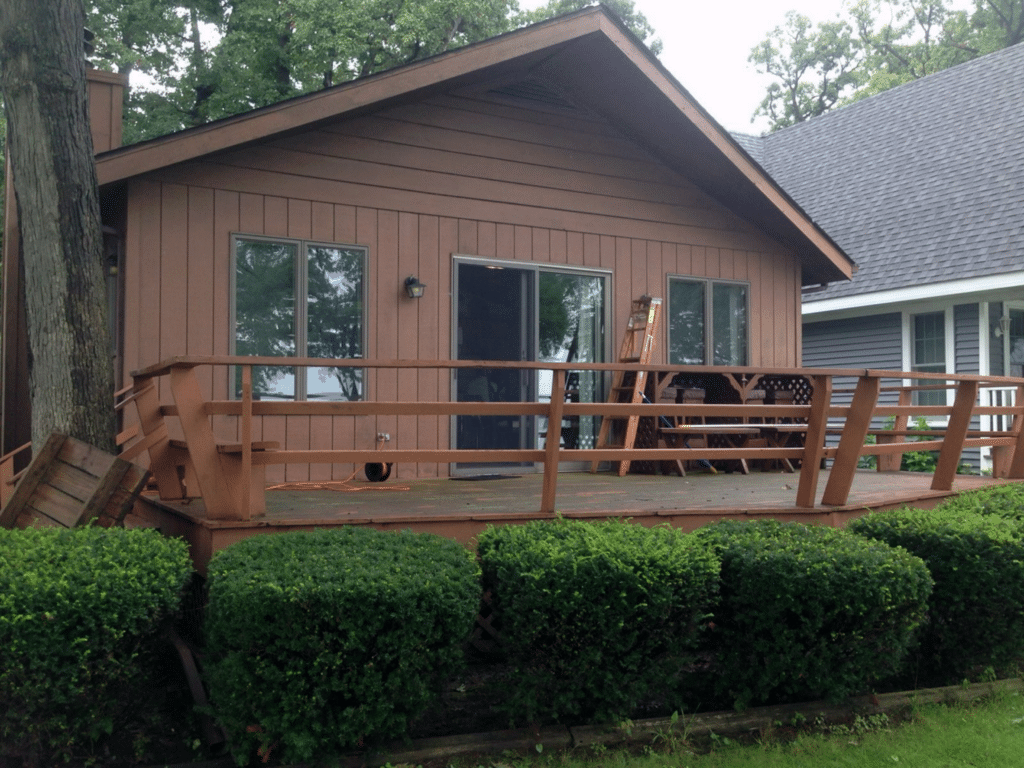 A brown wooden house with a damaged deck railing, green bushes in front, and a ladder leaning against the house.