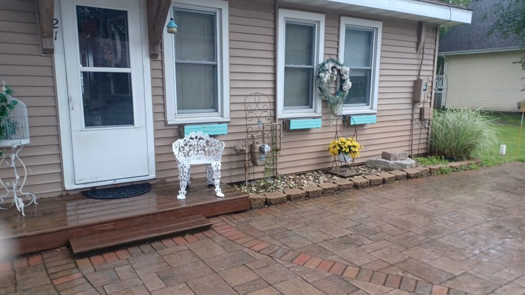 A paved patio leads to a beige house with two windows, a white chair, a wreath, yellow flowers, and decorative accents near the front door.