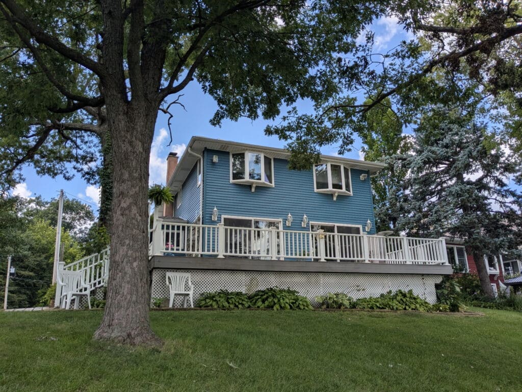 Two-story blue house with white trim and a large porch, surrounded by green lawn, trees, and shrubs on a sunny day.
