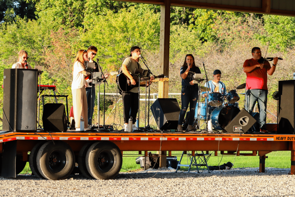 A band of seven people performs on an outdoor stage with instruments including keyboard, guitar, drums, violin, and microphones. Trees and sunlight are visible in the background.
