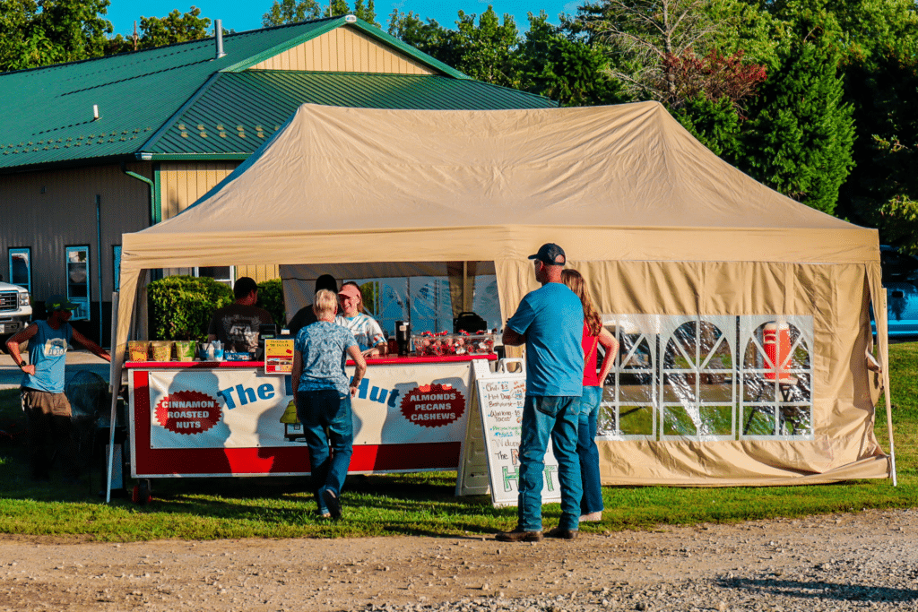 A tan canopy tent houses a vendor stand selling nuts at an outdoor fair. Several people are standing in front, and a menu board is displayed on the grass.